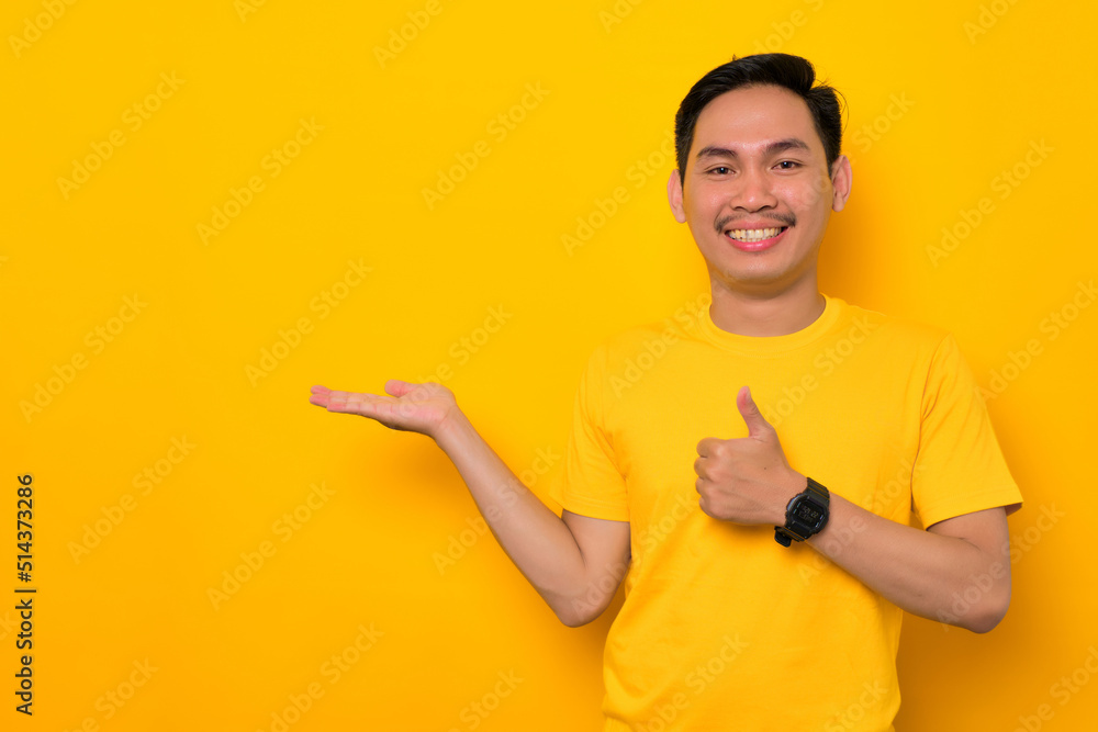 Cheerful young Asian man in casual t-shirt open palm showing thumbs up gesture isolated on yellow background. Great offer concept Cheerful young Asian man in casual t-shirt open palm showing thumbs up gesture isolated on yellow background. Great offer concept