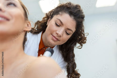 Dermatologist in latex gloves holding dermatoscope while examining attractive patient with skin disease. Female dermatologist examining patient with dermascope, looking for signs of skin cancer. photo
