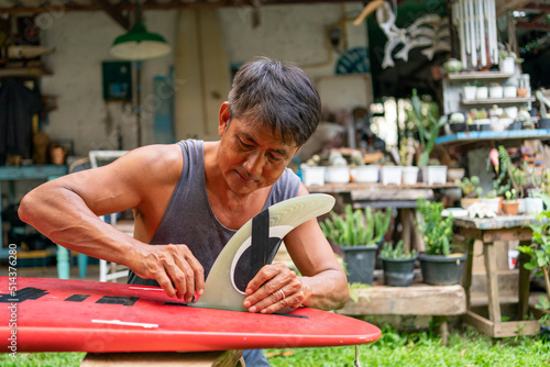Photography Asian senior man attaching fins on bottom of surfboard at home