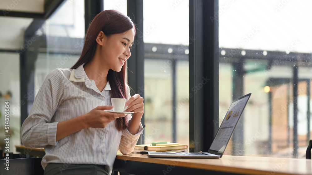 Relaxed young employee holding coffee cup and looking at screen of laptop while enjoying work in office