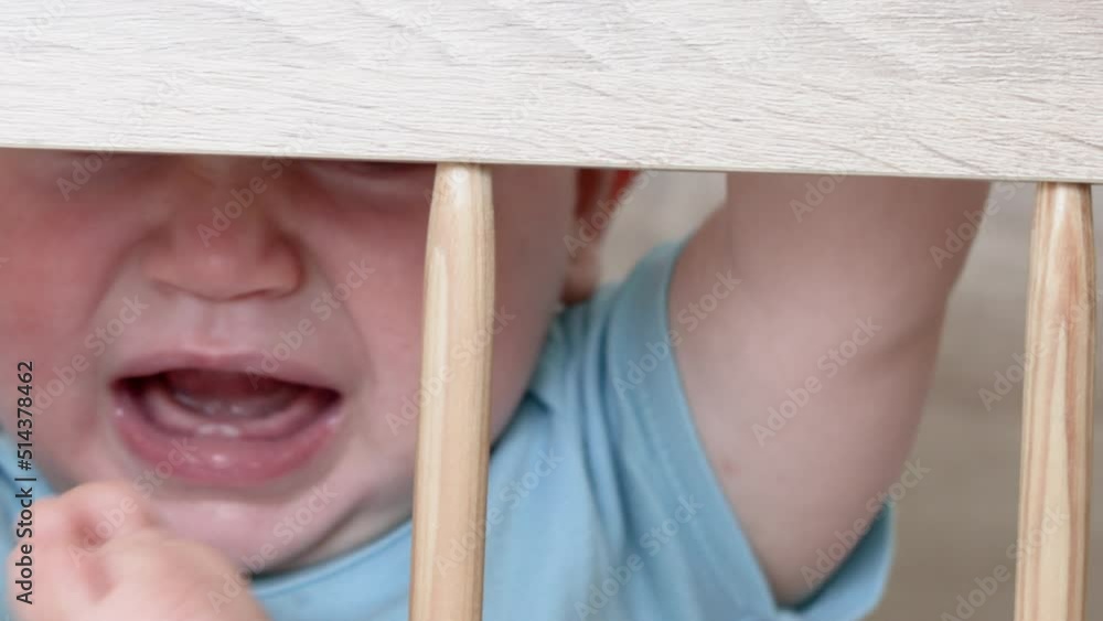 cute little baby boy is crying in the crib, holding the wooden bars