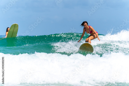 Asian woman surfer surfing and riding surfboard  the wave in the sea at tropical beach in sunny day. Healthy female enjoy outdoor activity lifestyle and water sport exercise surfing on summer vacation