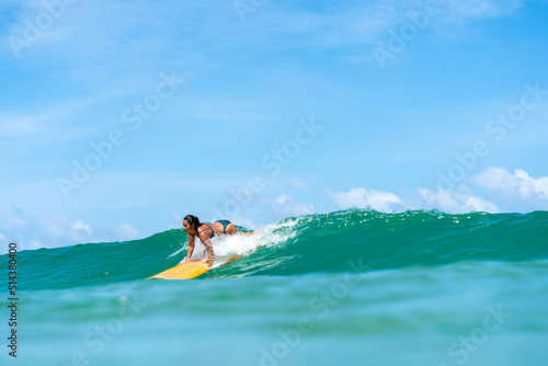 Asian woman surfer surfing and riding surfboard the wave in the sea at tropical beach in sunny day. Healthy female enjoy outdoor activity lifestyle and water sport exercise surfing on summer vacation