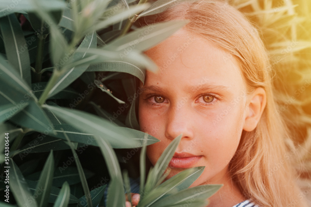 Naklejka premium portrait face of candid happy little kid girl of eight years old with long blond hair and green eyes on background of green plants during a summer vacation travel. gen z mental health concept. flare