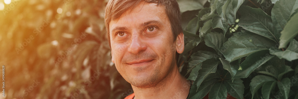 portrait of face happy smiling unshaven mature 40+ years man solo traveler with stubble on the background of green trees plants in nature on sunny summer day on vacation travel. banner. flare portrait of face happy smiling unshaven mature 40+ years man solo traveler with stubble on the background of green trees plants in nature on sunny summer day on vacation travel. banner. flare