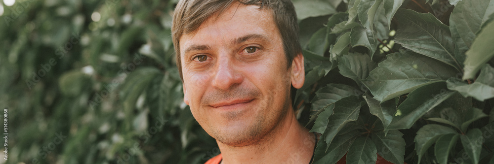 portrait of face happy smiling unshaven mature 40+ years man solo traveler with stubble and brown eyes on the background of green trees plants in nature on sunny summer day on vacation travel. banner portrait of face happy smiling unshaven mature 40+ years man solo traveler with stubble and brown eyes on the background of green trees plants in nature on sunny summer day on vacation travel. banner
