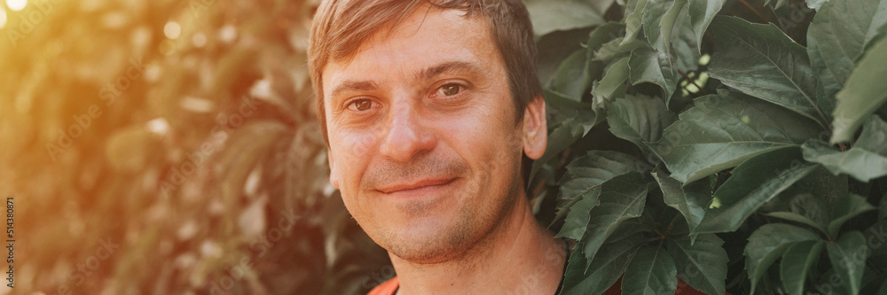 portrait of face happy smiling unshaven mature 40+ years man solo traveler with stubble on the background of green trees plants in nature on sunny summer day on vacation travel. banner. flare portrait of face happy smiling unshaven mature 40+ years man solo traveler with stubble on the background of green trees plants in nature on sunny summer day on vacation travel. banner. flare