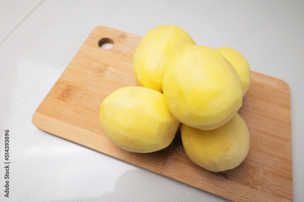 Peeled fresh potatoes lie on a wooden cutting board on a white table