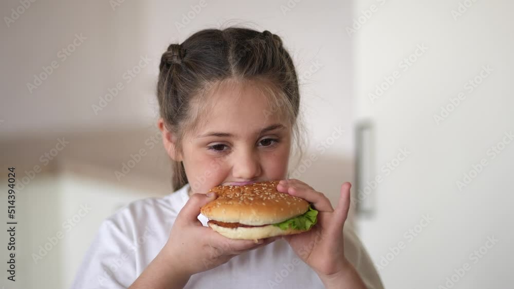 little girl eating a hamburger. unhealthy fast food proper nutrition concept. child greedily with pleasure bites a big burger in the kitchen at lifestyle home. kid eats fast food close-up little girl eating a hamburger. unhealthy fast food proper nutrition concept. child greedily with pleasure bites a big burger in the kitchen at lifestyle home. kid eats fast food close-up