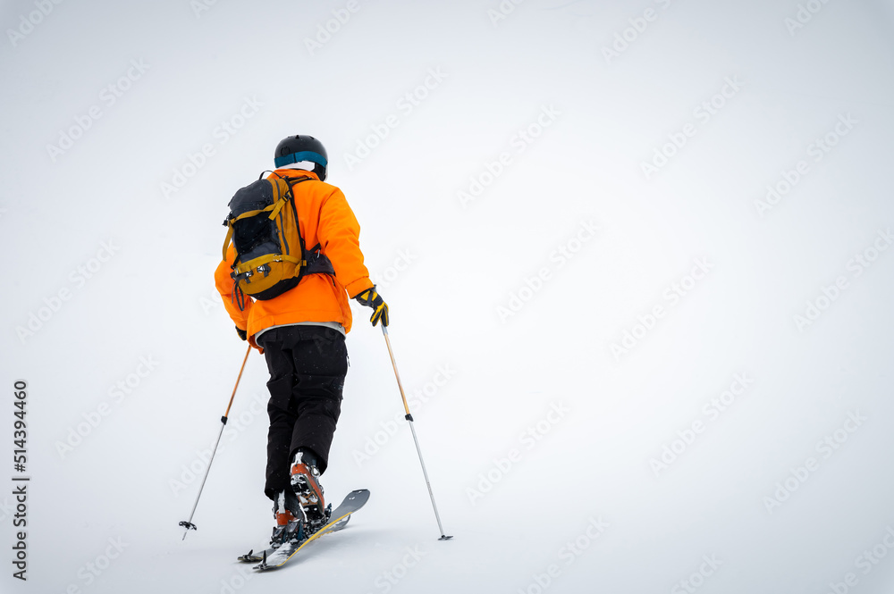 A male athlete in an orange jacket with a backpack on his shoulders and a helmet with a mask in a ski tour climbs a snowy slope in foggy weather. Ski touring and skiing copy space A male athlete in an orange jacket with a backpack on his shoulders and a helmet with a mask in a ski tour climbs a snowy slope in foggy weather. Ski touring and skiing copy space