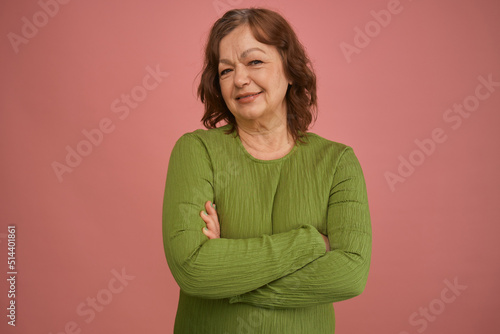 Portrait of a cute, optimistic, smiling, sweet senior mature woman with a kind expression on her face in a green dress coquettishly looking at the camera. Isolated on a pink background