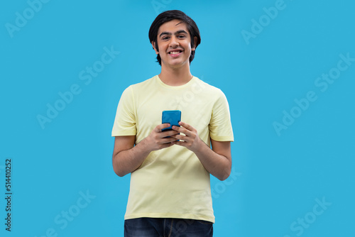 Portrait of a happy teenage boy using Smartphone while standing against blue background