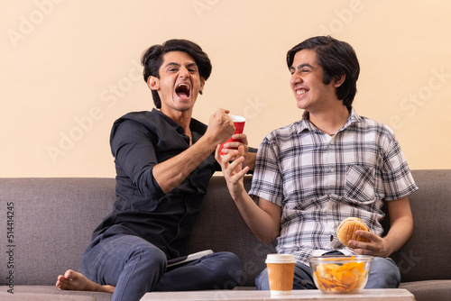 Two happy teenage boys eating fast foods and watching TV together at home