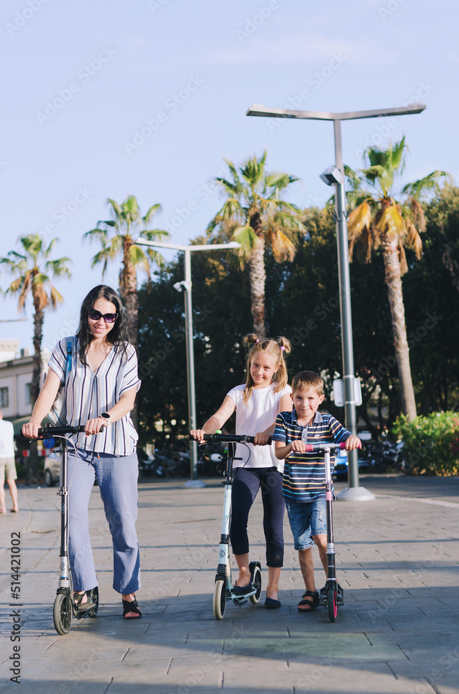 Fototapeta premium Happy family lifestyle and holiday concept. Mother, little boy, girl , riding scooters, walking in old city, street. Laughing on a summer sunny day. Having fun