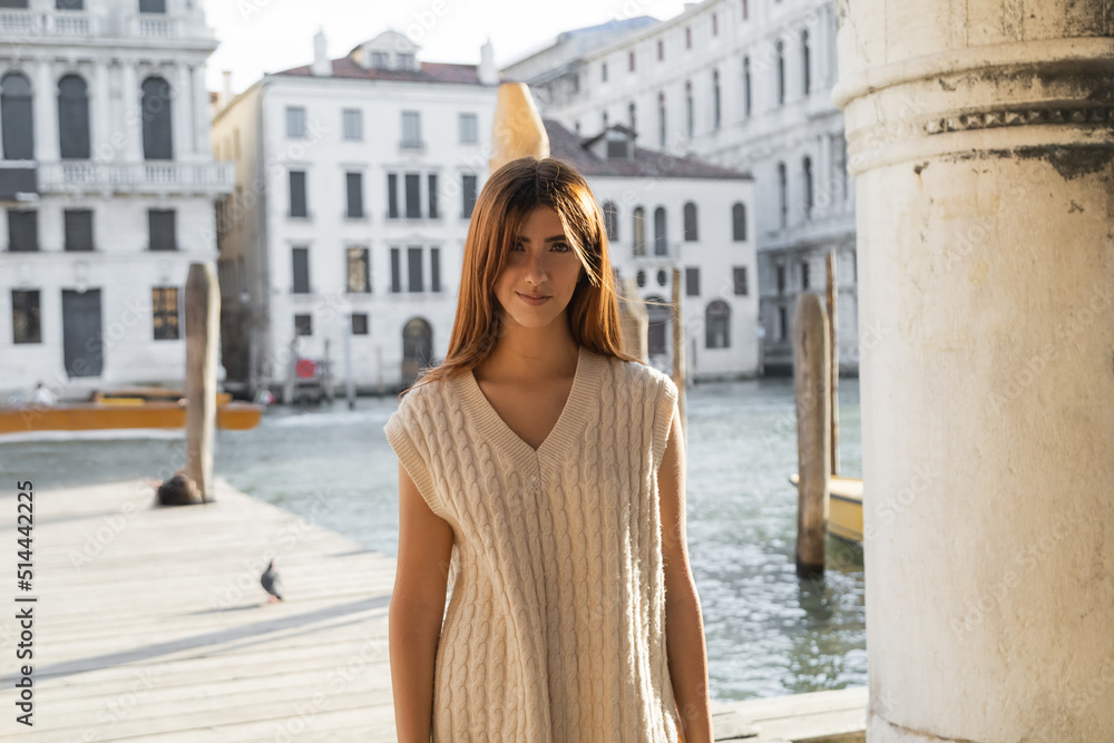 Fototapeta premium redhead woman in sleeveless jumper looking at camera near blurred Grand Canal in Venice.