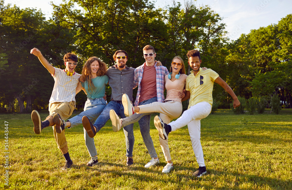 Funny group of friends having fun and fooling around dancing cancan in park on green lawn. Cheerful multiracial young people in casual clothes stand in row and laugh out loud, raising their legs. Funny group of friends having fun and fooling around dancing cancan in park on green lawn. Cheerful multiracial young people in casual clothes stand in row and laugh out loud, raising their legs.