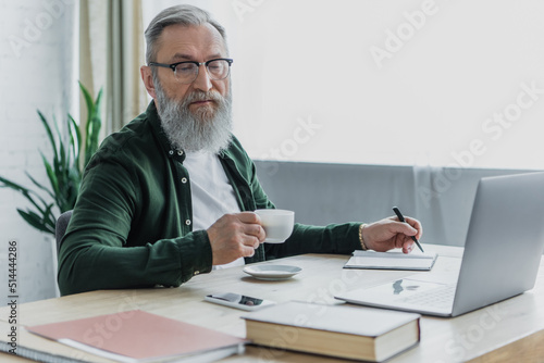 bearded senior man in eyeglasses holding cup of coffee and pen near laptop while working from home.