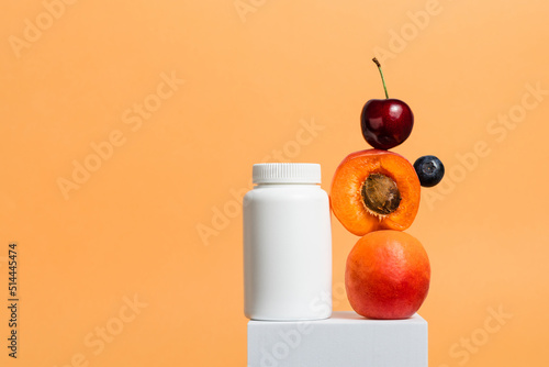 White bottle of multivitamin supplement with fresh fruits on cube stand, orange background, copy space photo