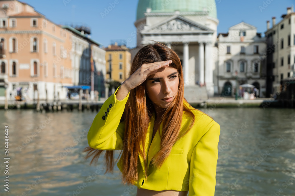trendy woman holding hand above eyes while looking away on blurred background in Venice.