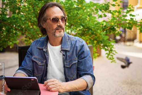 Middle aged man looking away sitting at cafe or street restaurant while working outdoors using digital tablet. Handsome mature freelancer man enjoying lunch outdoors while having group chat