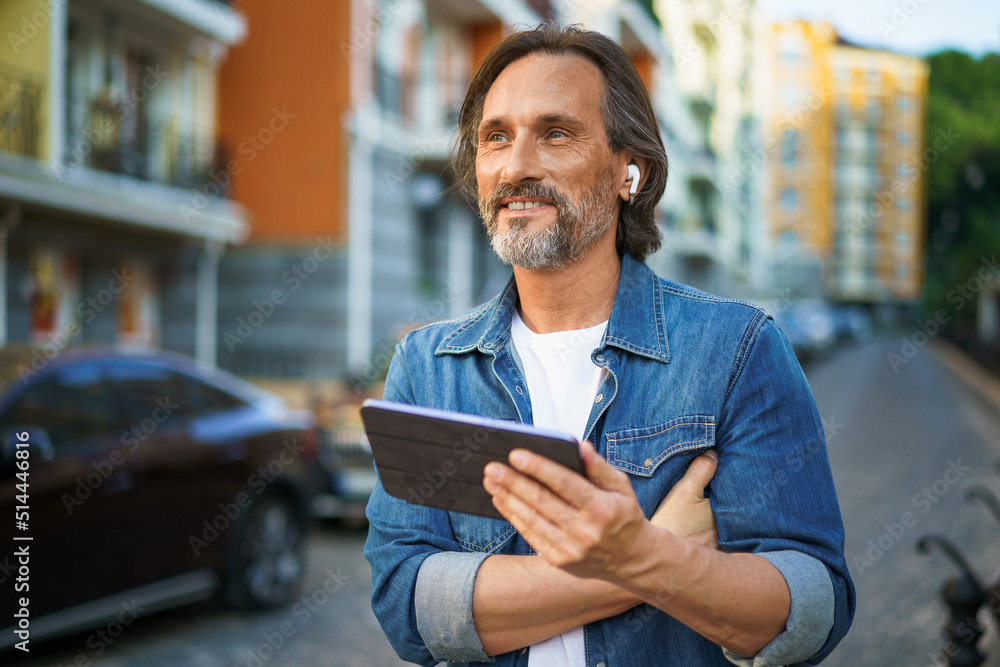 Handsome middle aged freelancer man using tablet having a call while walking on urban streets. Mature man use wireless earphones talking or listening music while travel in old town streets Handsome middle aged freelancer man using tablet having a call while walking on urban streets. Mature man use wireless earphones talking or listening music while travel in old town streets
