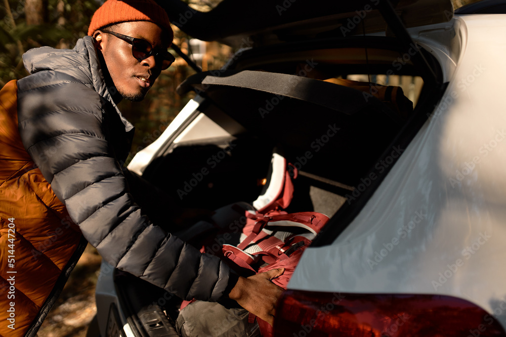 Fototapeta premium Outdoor closeup picture of african american man in spring coat, red hat and sunglasses putting things in car trunk, preparing to travel, for camping ride in mountains or in wild nature
