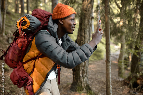 Side view of happy excited african american male hiker with backpack on shoulders walking in forest alone taking picture of wild plants, beauty of nature, dressed in spring clothes