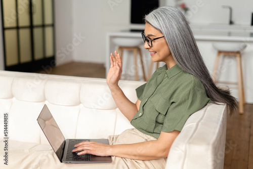 Video call concept. Happy senior woman using laptop computer for video connection from home, mature female looks at webcam and greeting colleagues, waving and smiles sitting on sofa at home