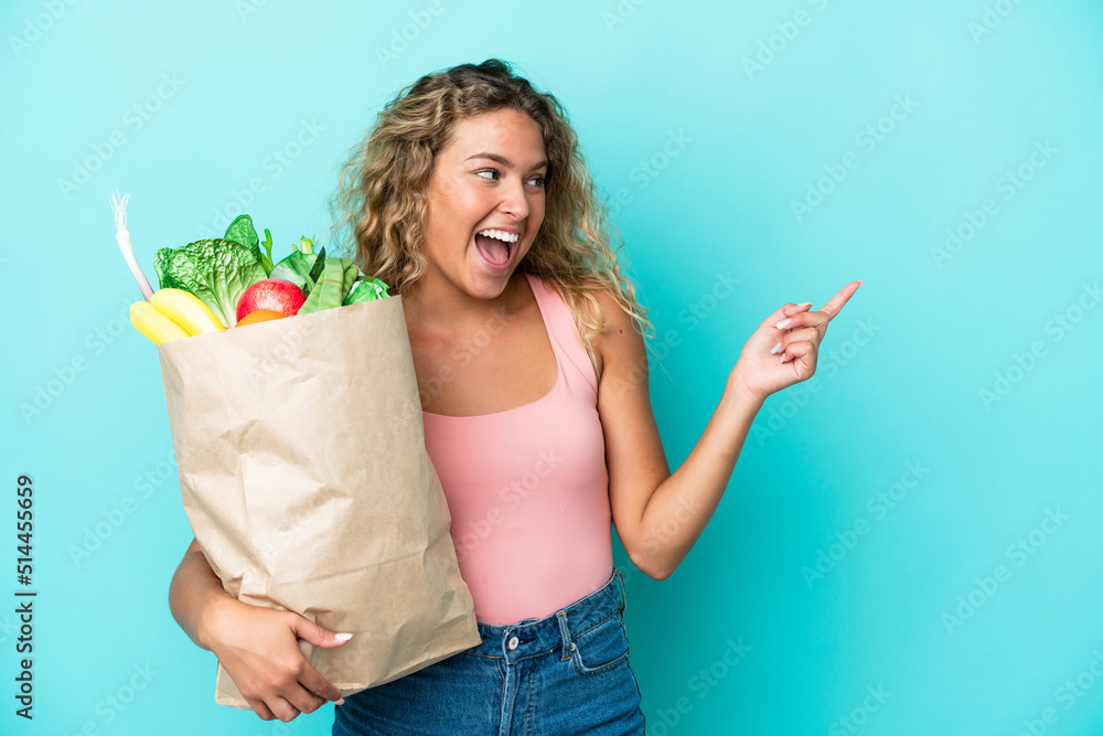 Girl with curly hair holding a grocery shopping bag isolated on green background pointing finger to the side and presenting a product Girl with curly hair holding a grocery shopping bag isolated on green background pointing finger to the side and presenting a product