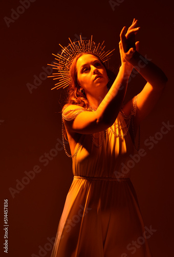 portrait of beautiful red-haired woman wearing long flowing fantasy toga gown with golden halo crown jewellery, creative hand gestures on a dark moody background with glowing orange lighting.