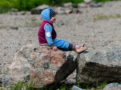 a little boy in a blue jumpsuit with a hood and a vest is sitting on huge rocks with his legs stretched out and laughing