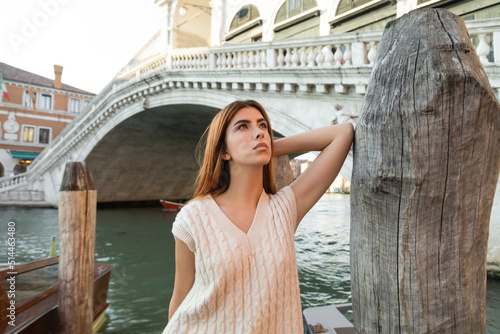 young woman looking away near wooden piling and Rialto Bridge on background in Venice.