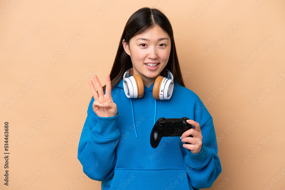 Young Chinese man playing with a video game controller isolated on beige background happy and counting three with fingers Young Chinese man playing with a video game controller isolated on beige background happy and counting three with fingers