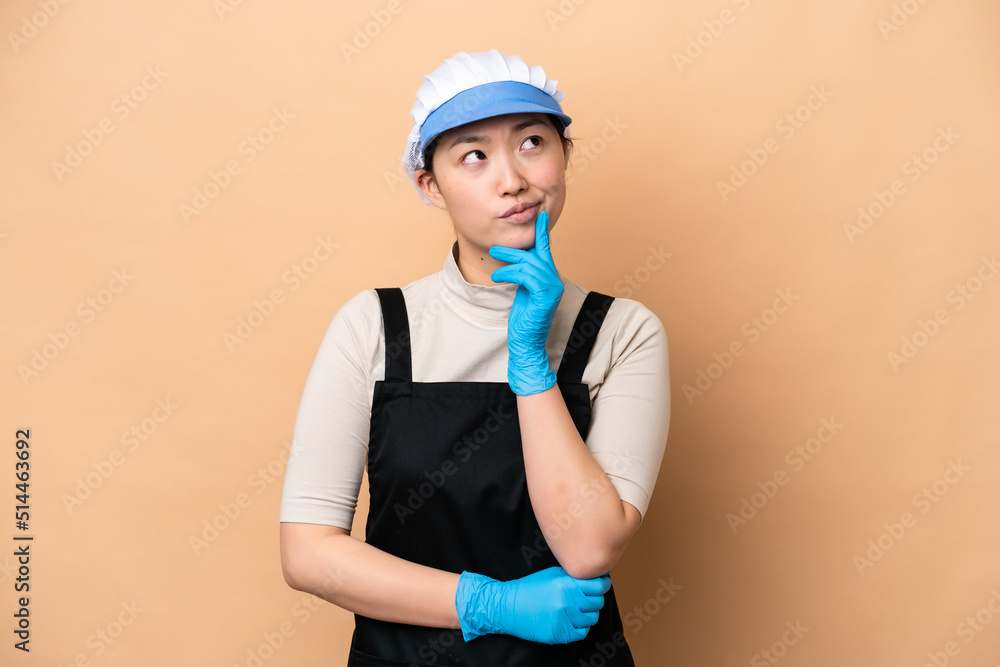 Young Chinese Fishmonger woman wearing an apron and holding a raw fish isolated on pink background having doubts while looking up
