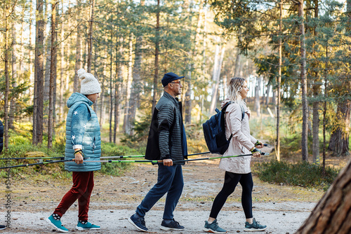 Three people with backpack make Scandinavian walking, hold trekking sticks as team group in forest. Exercise education