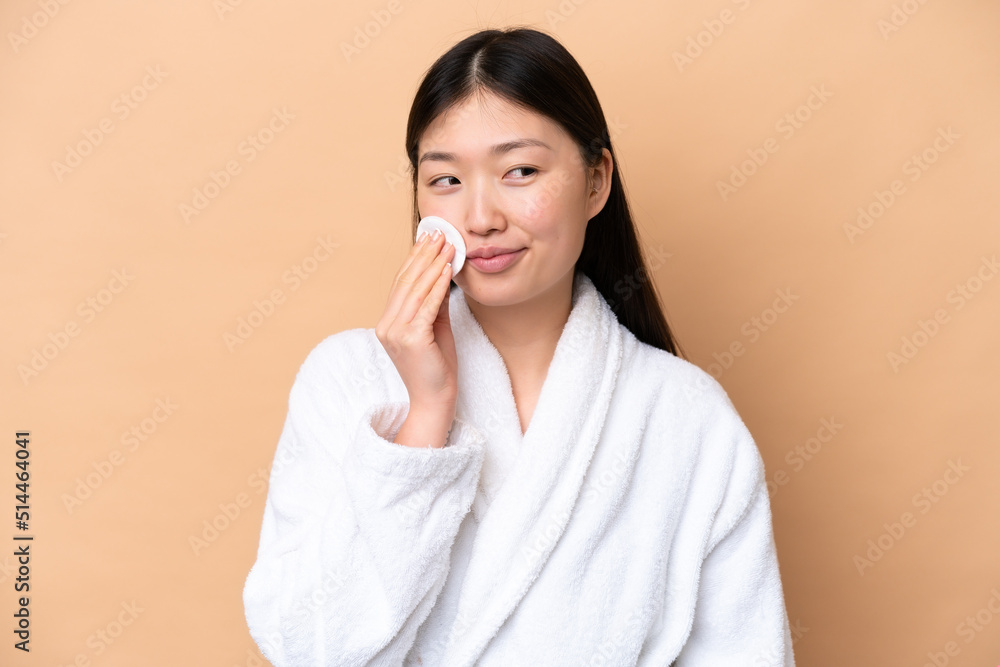 Young Chinese woman isolated on beige background with cotton pad for removing makeup from her face Young Chinese woman isolated on beige background with cotton pad for removing makeup from her face