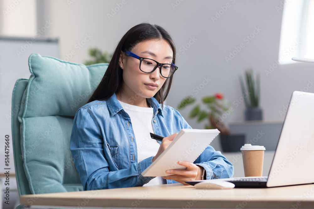 Young Asian woman studying online while sitting in office, writing lecture notes in notebook, using laptop for studying Young Asian woman studying online while sitting in office, writing lecture notes in notebook, using laptop for studying