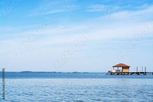 Sunny summer day with a cloudless blue sky, a wooden lonely house on the water of a lake or river, a horizontal photo without people