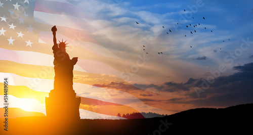 Statue of Liberty with a large american flag and sunset sky with flying birds on background.