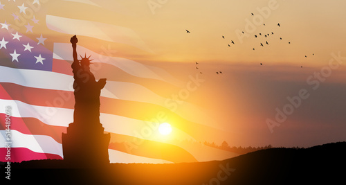Statue of Liberty with a large american flag and sunset sky with flying birds on background.