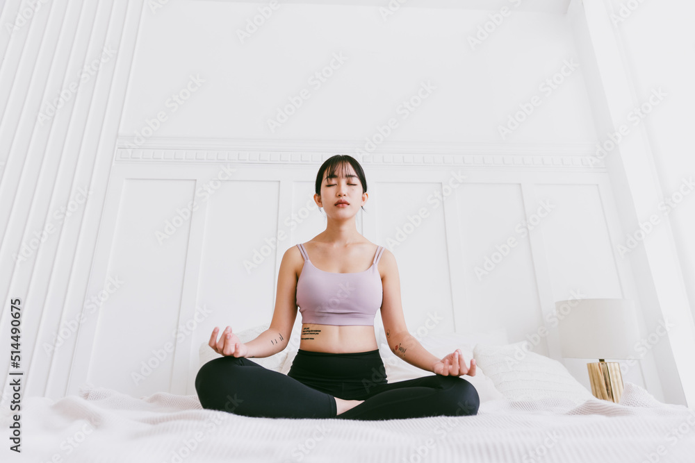 Fototapeta premium Sporty Asian woman practicing yoga on a white bed, doing Ardha Padmasana exercise, meditating in Half Lotus pose, indoor working out at home, wearing sportswear.