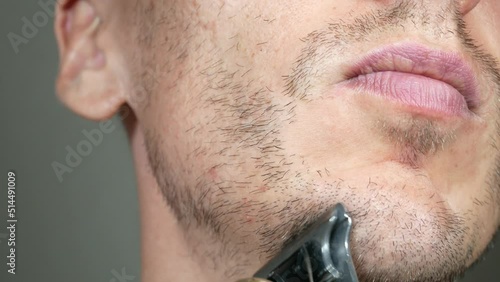 Man is cutting beard by Electric metal razor or dry shaver. Trimmer for cutting. Short, sparse beard on mans face. Hair growth problems. Unshaven bristles on the beard.