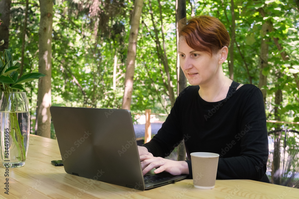 Woman works on a laptop while sitting at a wooden table in the yard of her country house. The concept of remote work. Focused woman solves work tasks on her day off. Woman works on a laptop while sitting at a wooden table in the yard of her country house. The concept of remote work. Focused woman solves work tasks on her day off.