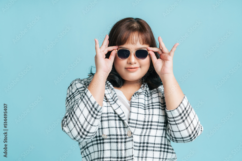 Portrait beautiful confidence Asian woman plus size model wearing sunglasses on isolate light blue studio background. Fashion young female chubby confident concept. Portrait beautiful confidence Asian woman plus size model wearing sunglasses on isolate light blue studio background. Fashion young female chubby confident concept.