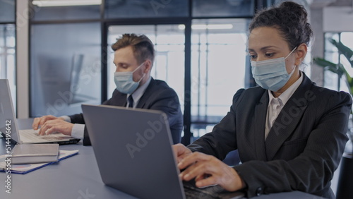 Two colleagues working on laptops in office, wearing face masks, pandemic safety