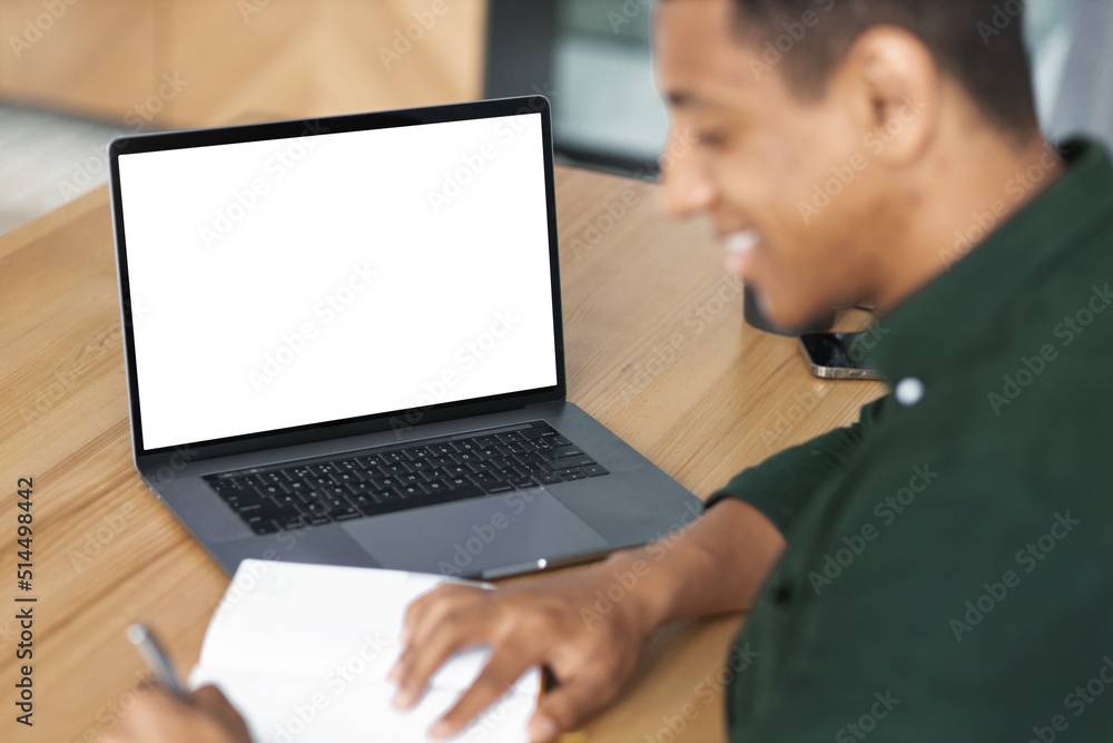 African American young student or freelancer man working remotely, takes notes, communicates online using video call sitting at workplace with laptop with blank screen, mockup