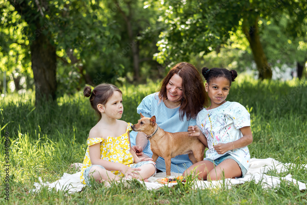 A multiracial family with a dog is having a picnic on a summer day.Mother and two daughters playing with the dog and having fun.Family, summer,diverse people concept. A multiracial family with a dog is having a picnic on a summer day.Mother and two daughters playing with the dog and having fun.Family, summer,diverse people concept.