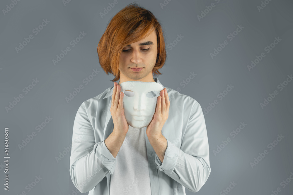 Mental health concept. Young man holding a full-face mask, ball, or masquerade carnival mask. Symbol of bipolar disorder or hypocrisy, deceit, duplicity. Portrait of guy Prep Student with long hair