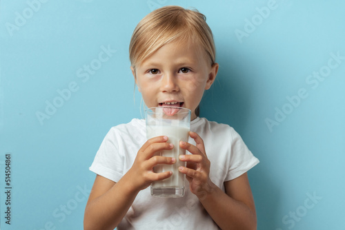 Cute toddler caucasian girl smiling and drinking milk from the glass on the blue background. Benefits of milk and calcium for children