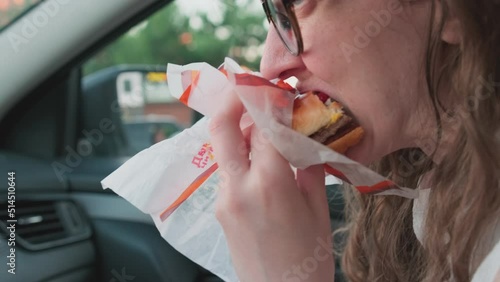 Woman with fast food inside of car. Woman holding burger biting it, eating and savouring food on passenger seat of a car. Fast snack on the go